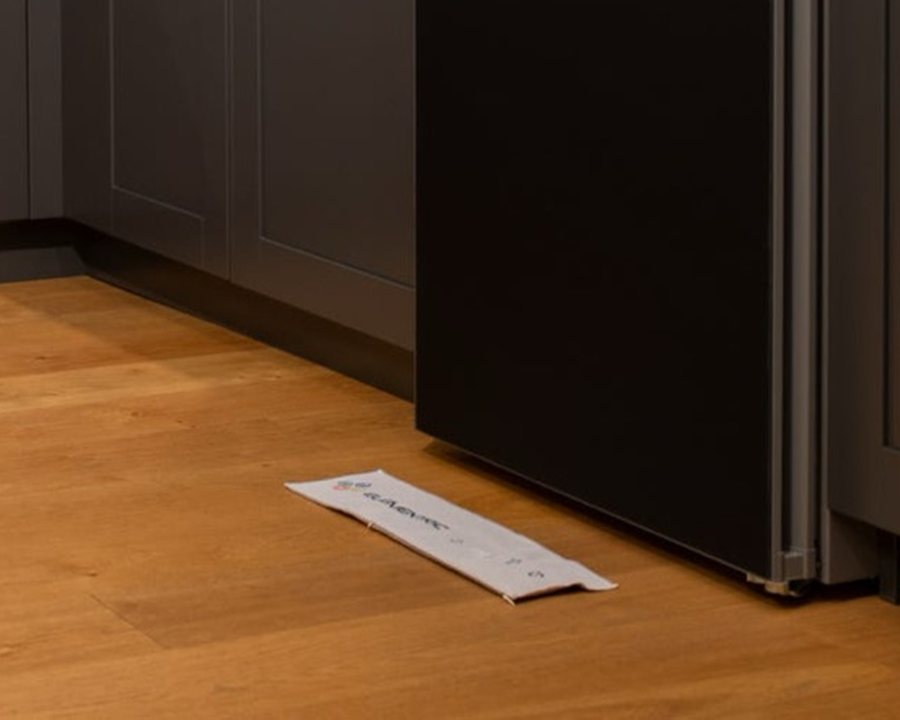 A white Dripspy mat on a wooden kitchen floor, beside the base of a tall fridge